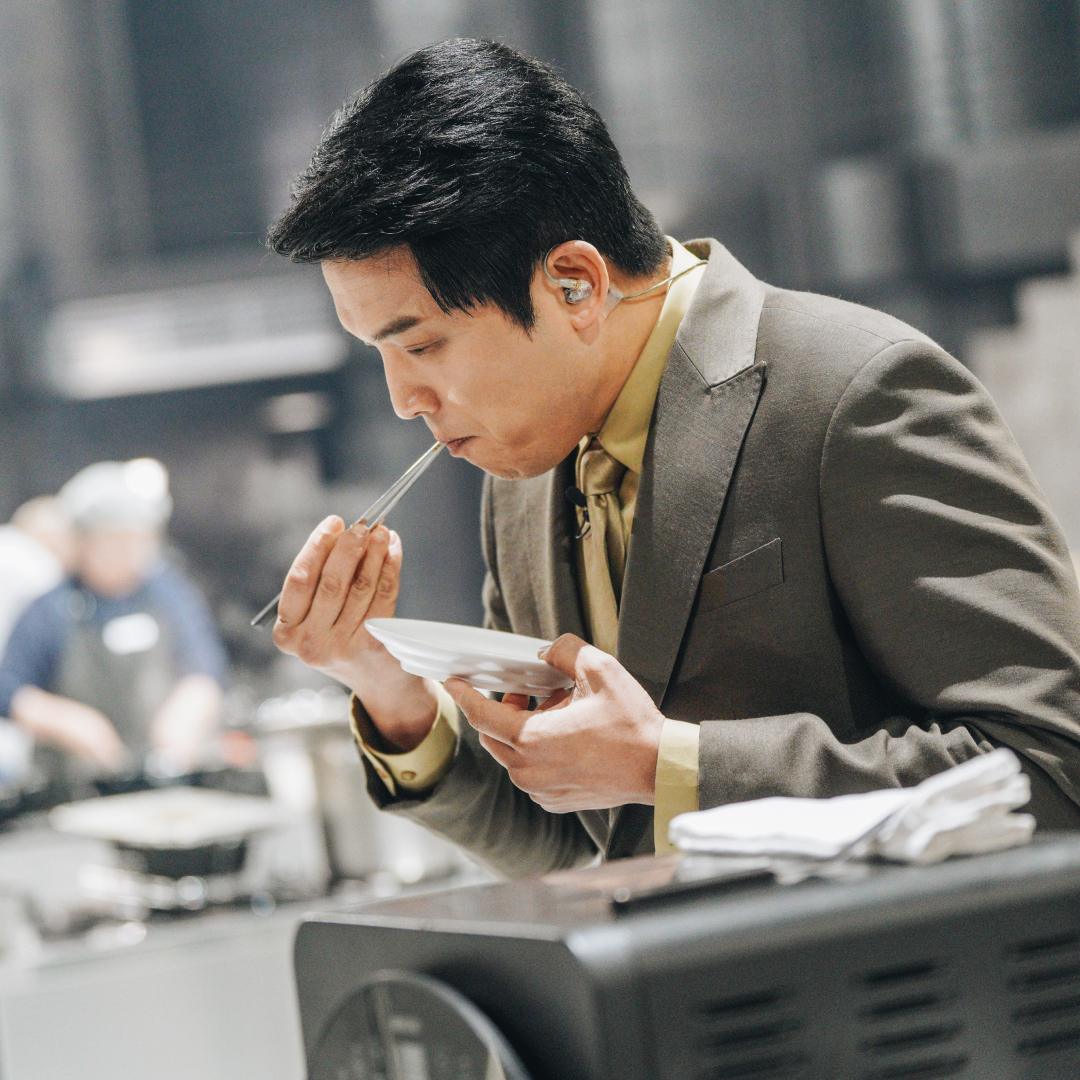 A Korean man (chef Ahn Sung-jae) tastes a meal with chopsticks. Chefs work at cook stations the blurred background, in a still from Netflix's 'Culinary Class Wars.'