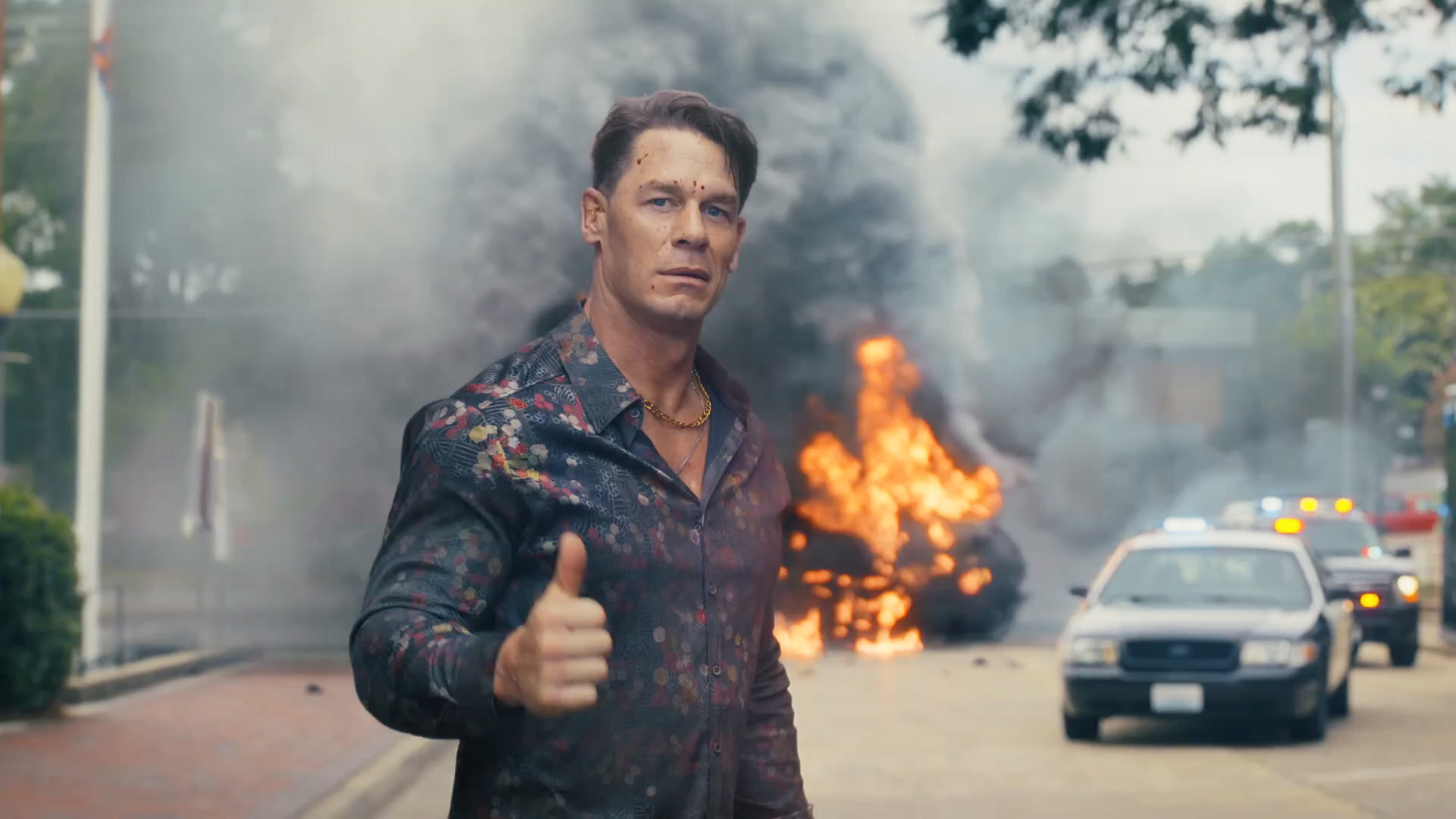 John Cena's Chris Smith smiling and giving a thumbs up as a car explodes behind him in Peacemaker season 2