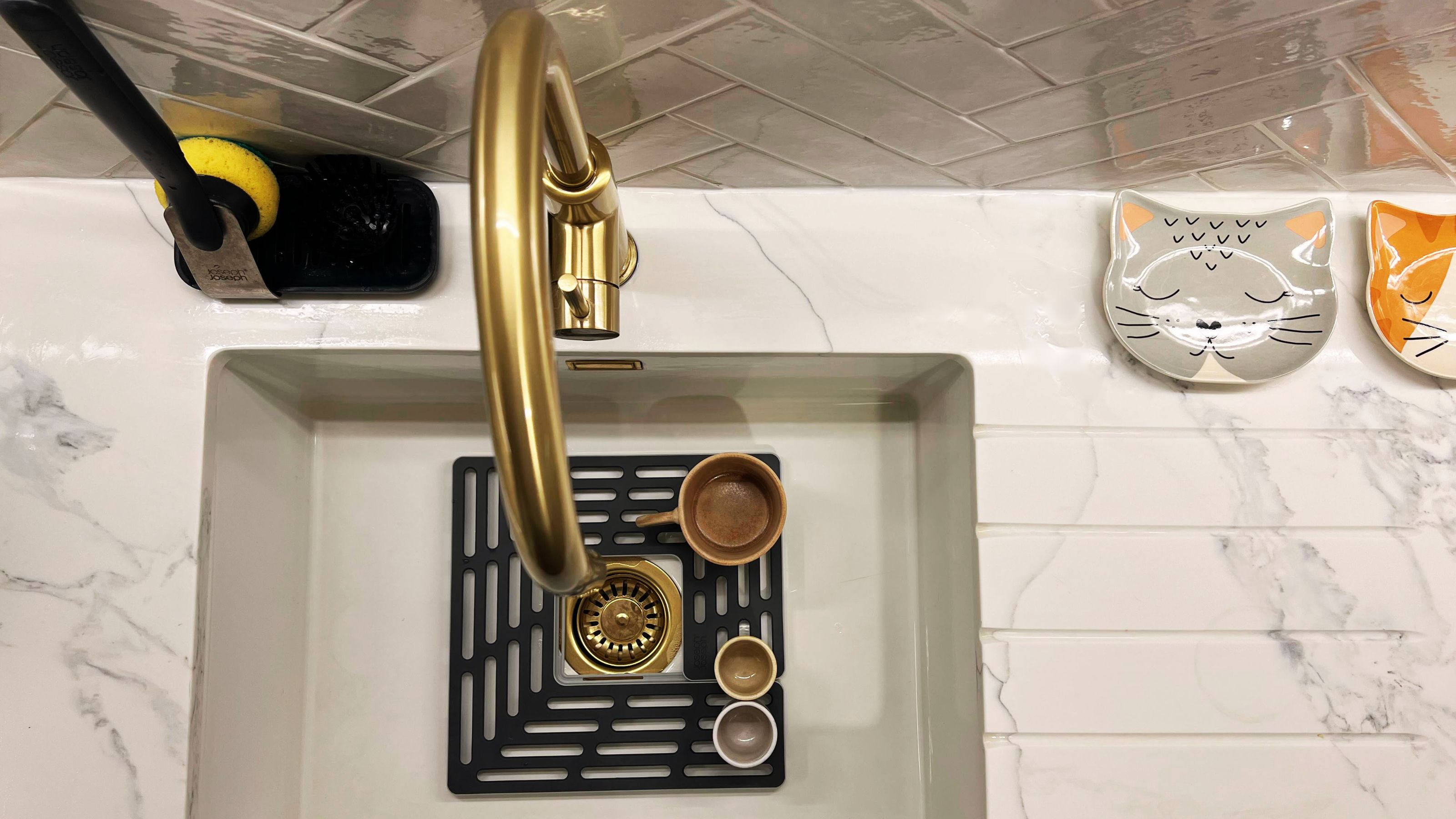 A kitchen sink with a sink protector inside as well as a washing sponge and a view of a cat feeding dish on the side of the kitchen counter