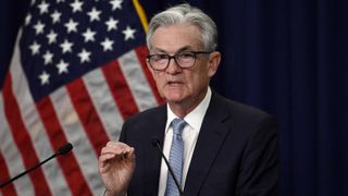 Fed Chair Jerome Powell gestures as he speaks into a microphone at a press conference, with an American flag in the background behind him.