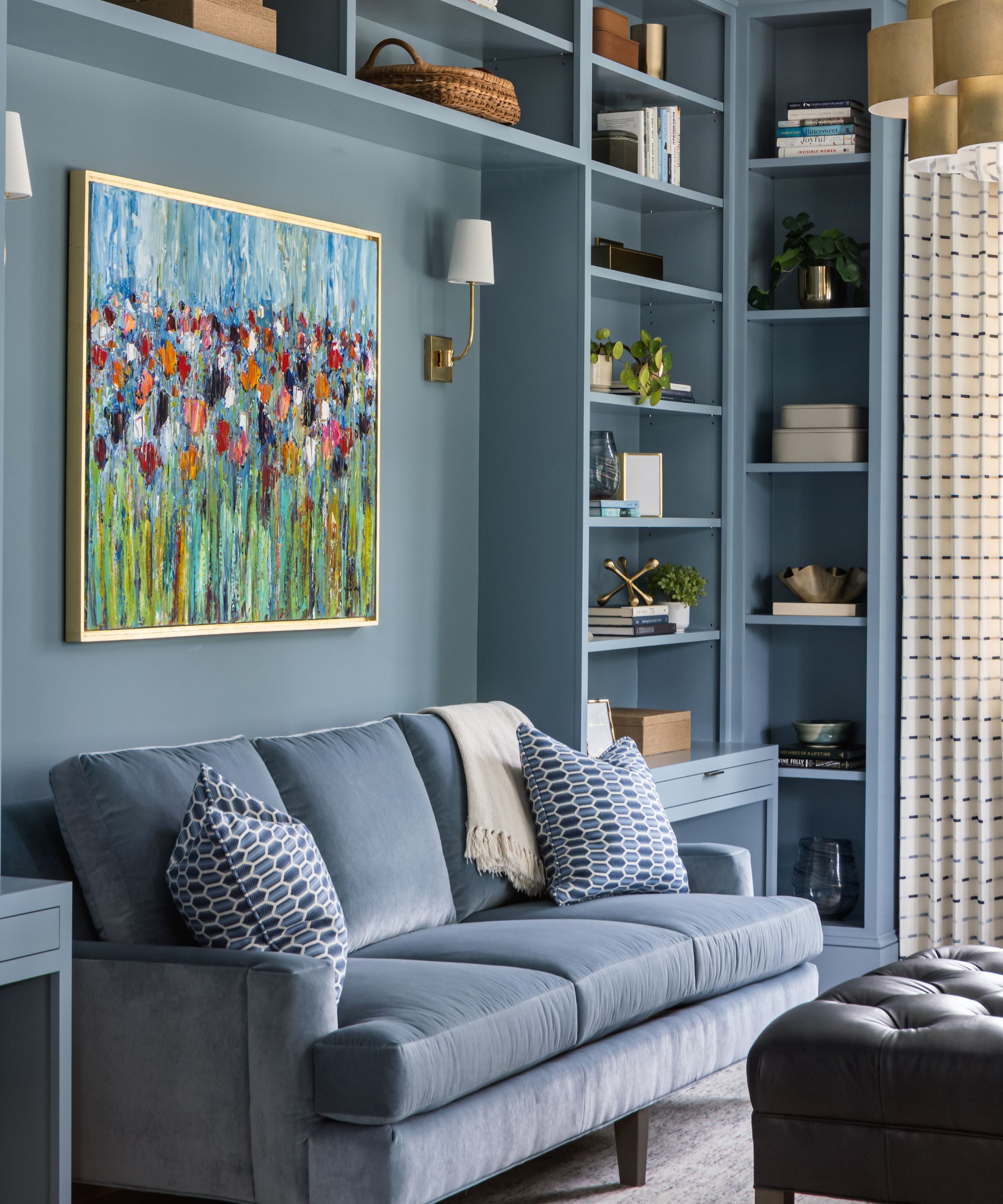 A home library room with muted blue color-drenched walls and built-in shelving. A dusky blue sofa with blue cushions, and a large piece of floral artwork on the wall with two wall sconces.