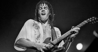 A black and white image of an open-shirted Pat Travers playing a Gibson double cut onstage in 1978