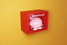 Pink piggy bank in a red box with a glass printed with the instruction BREAK GLASS IN CASE OF EMERGENCY on a yellow wall.