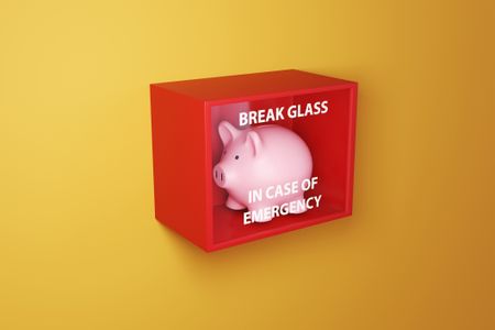 Pink piggy bank in a red box with a glass printed with the instruction BREAK GLASS IN CASE OF EMERGENCY on a yellow wall.