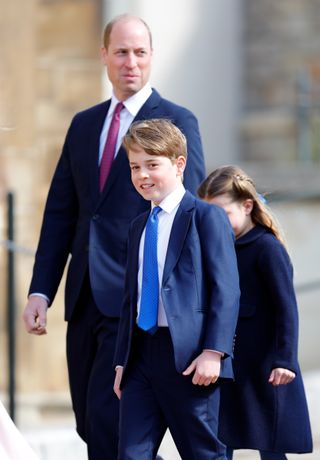 Prince George wears a navy suit with a white shirt and blue tie and is followed by dad Prince William