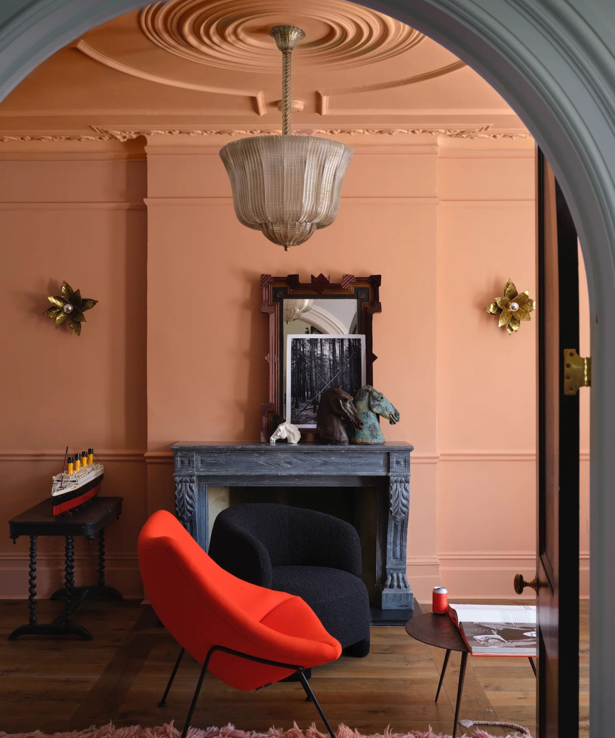 A vibrant room with peach-colored walls and ceiling, featuring an ornate grey fireplace, a modern red lounge chair, and a black textured armchair under a glass chandelier.