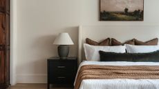 A white bedroom with a black nightstand, and a table lamp, a framed painting, a white bed, an auburn coverlet, and pillows