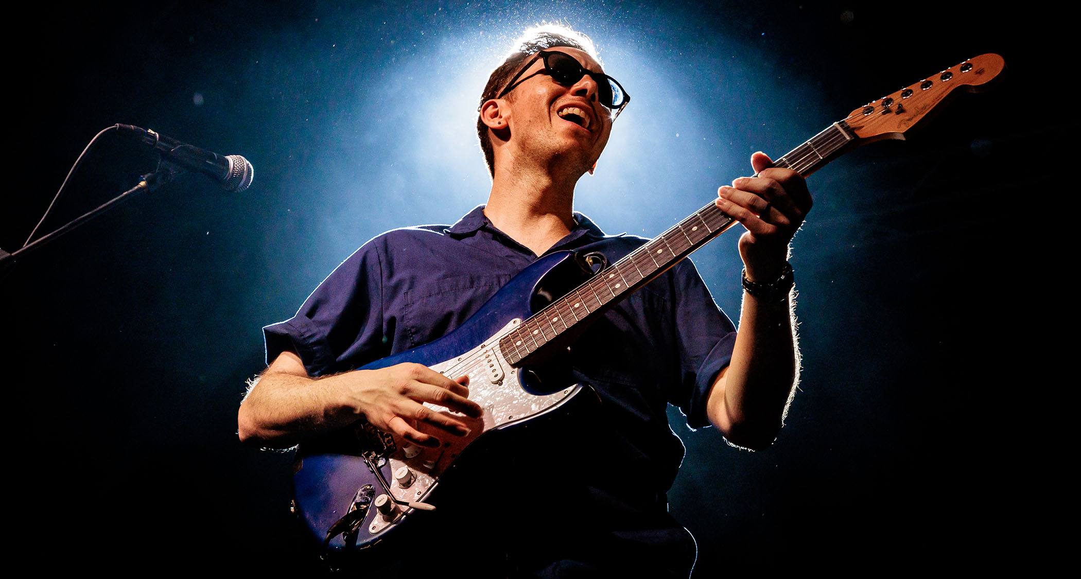 Cory Wong wears shades and performs with his blue Strat live in Italy, 2025, and is lit from behind by a white stage light.