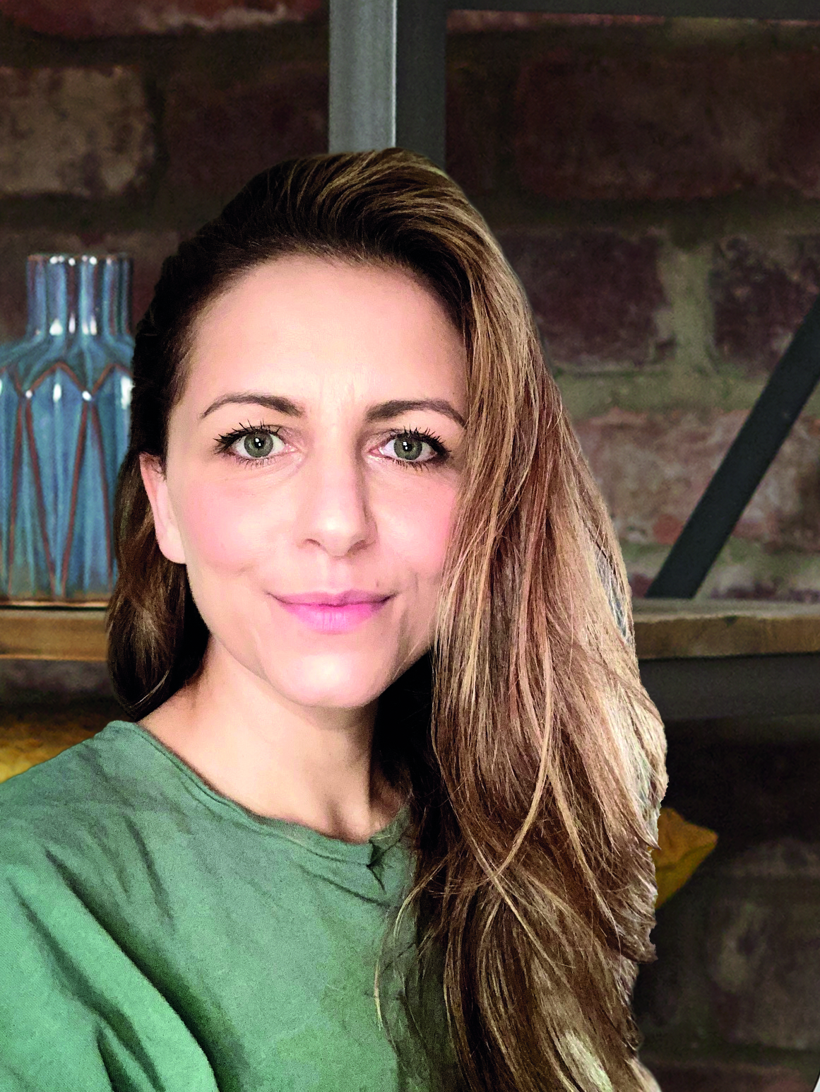 woman with long brown hair smiles to camera with a brick wall behind