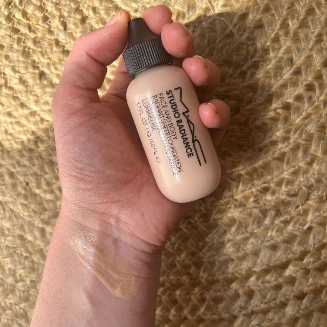 Beauty Editor Fiona McKim's hand holding a tube of foundation, with a swatch of the foundation on her wrist, on a woven jute rug backdrop