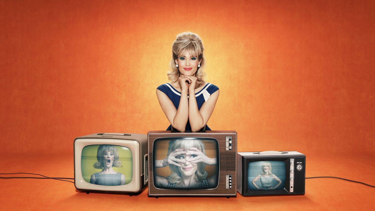 Funny Woman – Gemma Arterton in a blue and white dress and blonde wig as Barbara Parker, leans on 3 TVs which all have shots of her in comic poses