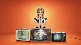 Funny Woman – Gemma Arterton in a blue and white dress and blonde wig as Barbara Parker, leans on 3 TVs which all have shots of her in comic poses