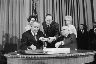 President Lyndon B. Johnson signs the Medicare bill as Vice President Humphrey and Harry S. Truman both check the time.