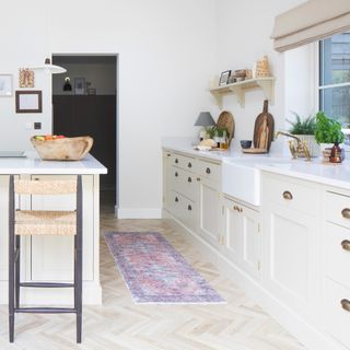country cottage kitchen with cream coloured shaker cabinets and cup handles, a kitchen island with a wicker bar stool and a patterned runner on timber herringbone flooring