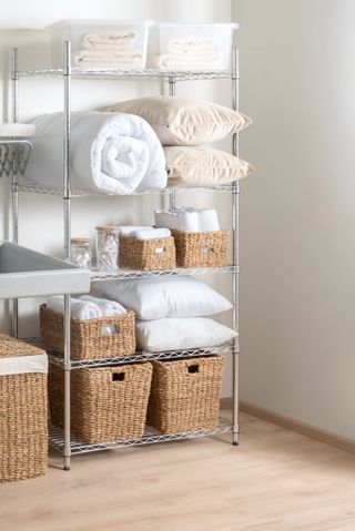 Interior of Bright Modern Laundry Room with White Duvet, Towels, Cozy Pillows, and Wicker Baskets on Adjustable Metal Storage Rack with Shelves, Chrome Wire Shelving Unit, Over Wooden Flooring.