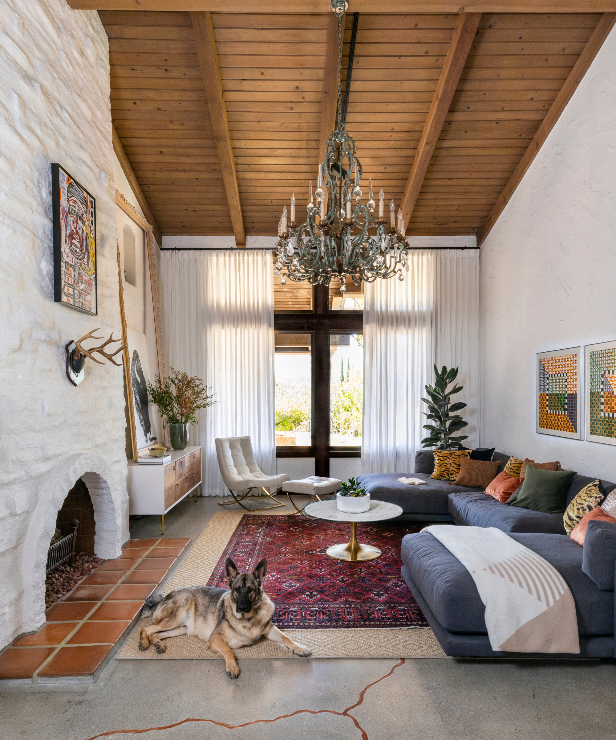 a living room with a vaulted, wood-paneled ceiling, a large blue/gray couch with several pillows, white walls, floor to ceiling windows covered by white drapes in the background, a large chandelier hanging from the ceiling, and a persian rug on top of a jute rug on the floor
