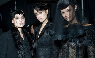 3 models backstage wearing all black formal attire with various different headwear