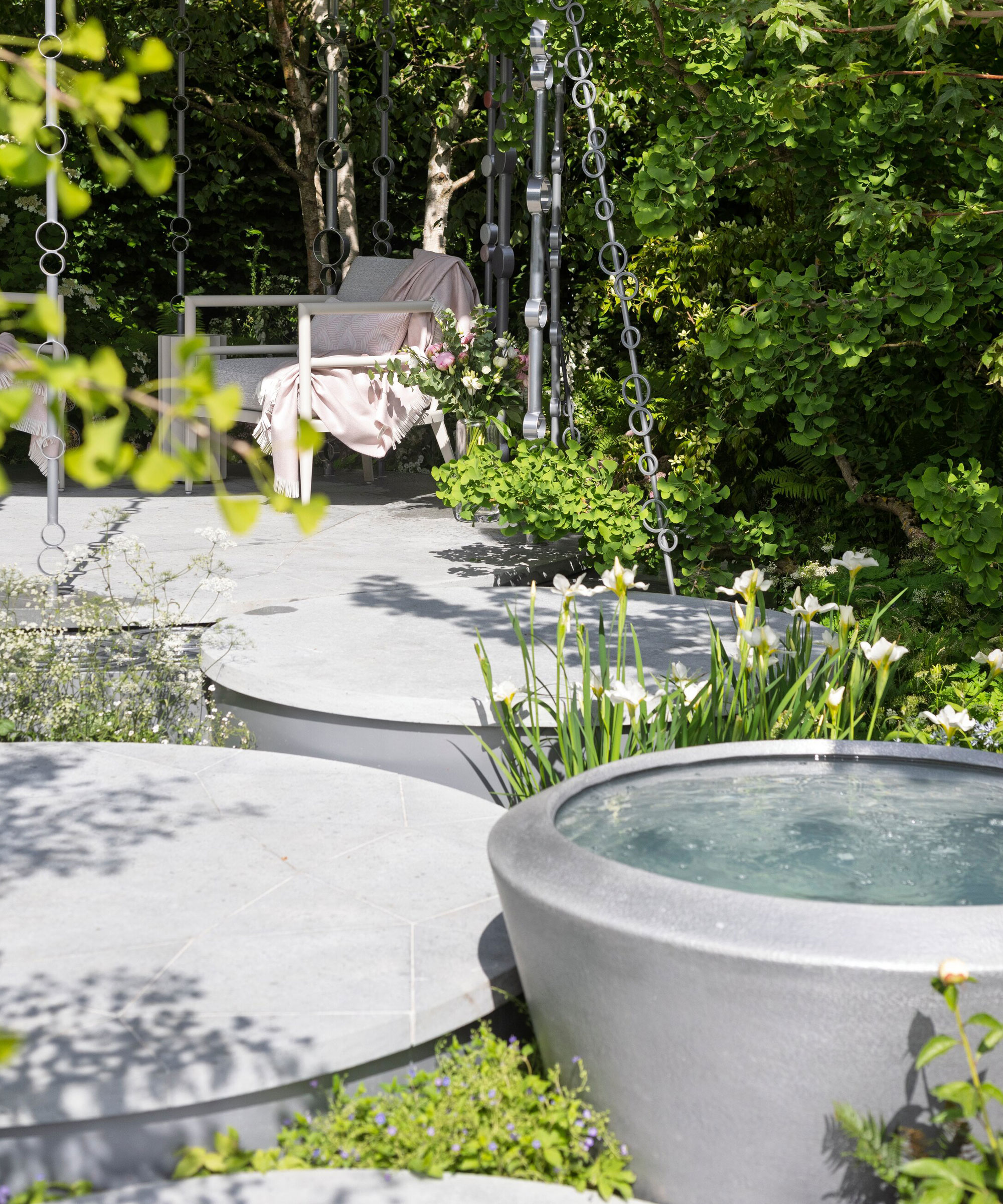 raised circular water feature with small circular patios and garden chair, surrounded by planting and shrubs