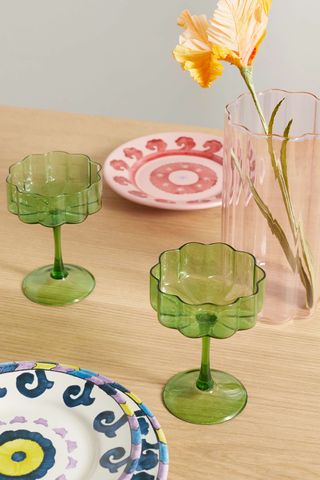 two green wavy-edged cocktail glasses on a table with colorful plates and flowers