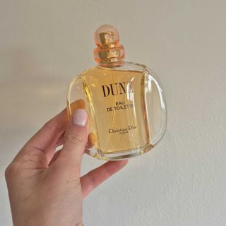 A close-up of Digital Beauty Writer, Naomi Jamieson's hand holding up Dior's Dune perfume against a cream wall