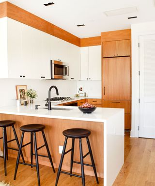 A white small kitchen with light wooden cupboards, white cabinets with a silver microwave within it, a sink, and a white rectangular kitchen island with three black circular wooden stools underneath it