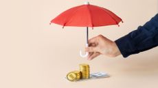 Hand holding a red umbrella over a stack of coins and notes on a cream-colored plain background.