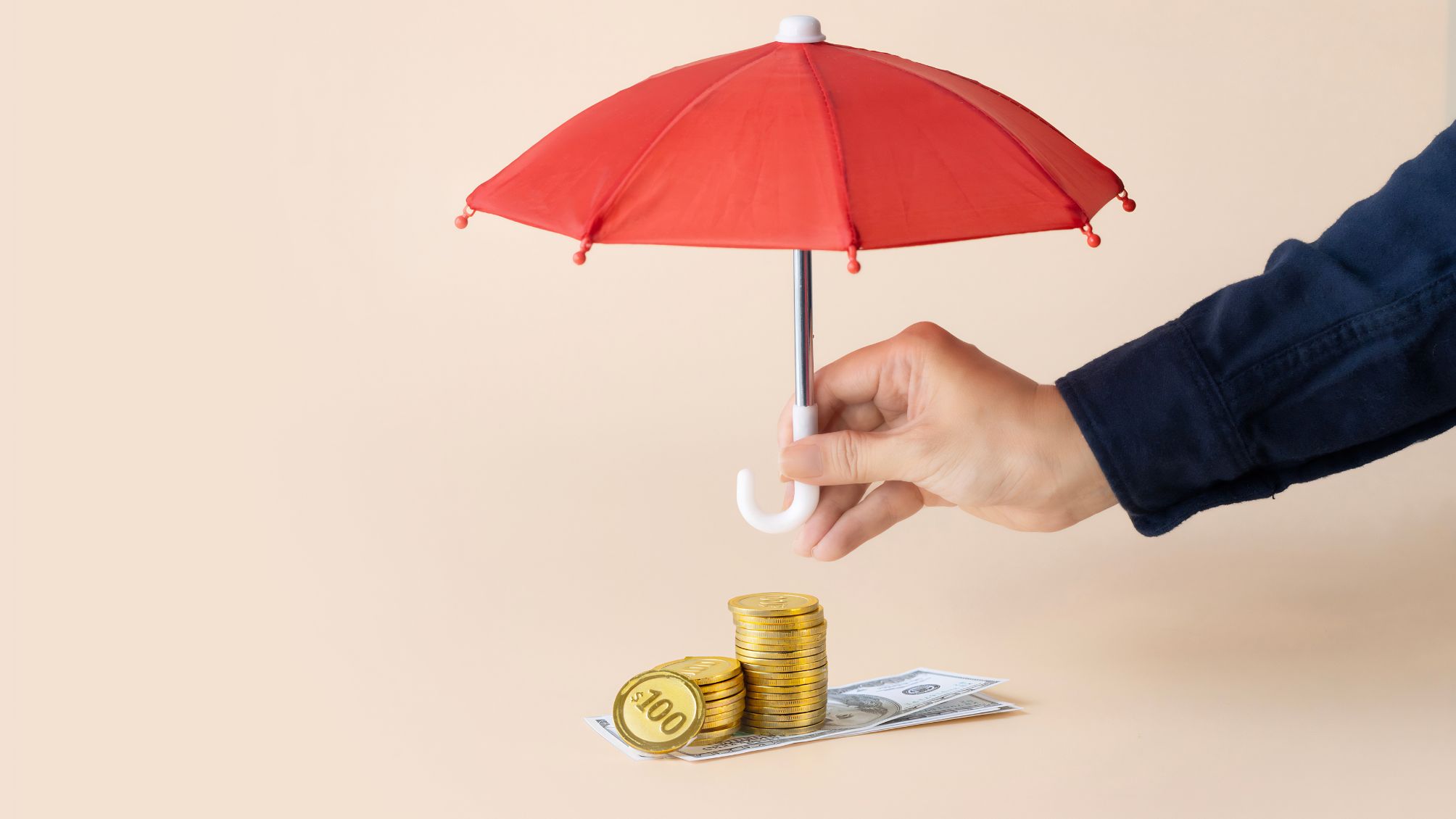 Hand holding a red umbrella over a stack of coins and notes on a cream-colored plain background.
