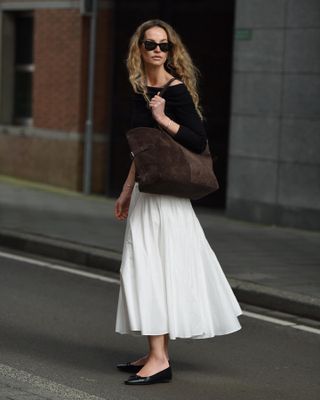 Woman wearing white maxi skirt, black sweater, and large suede tote bag.