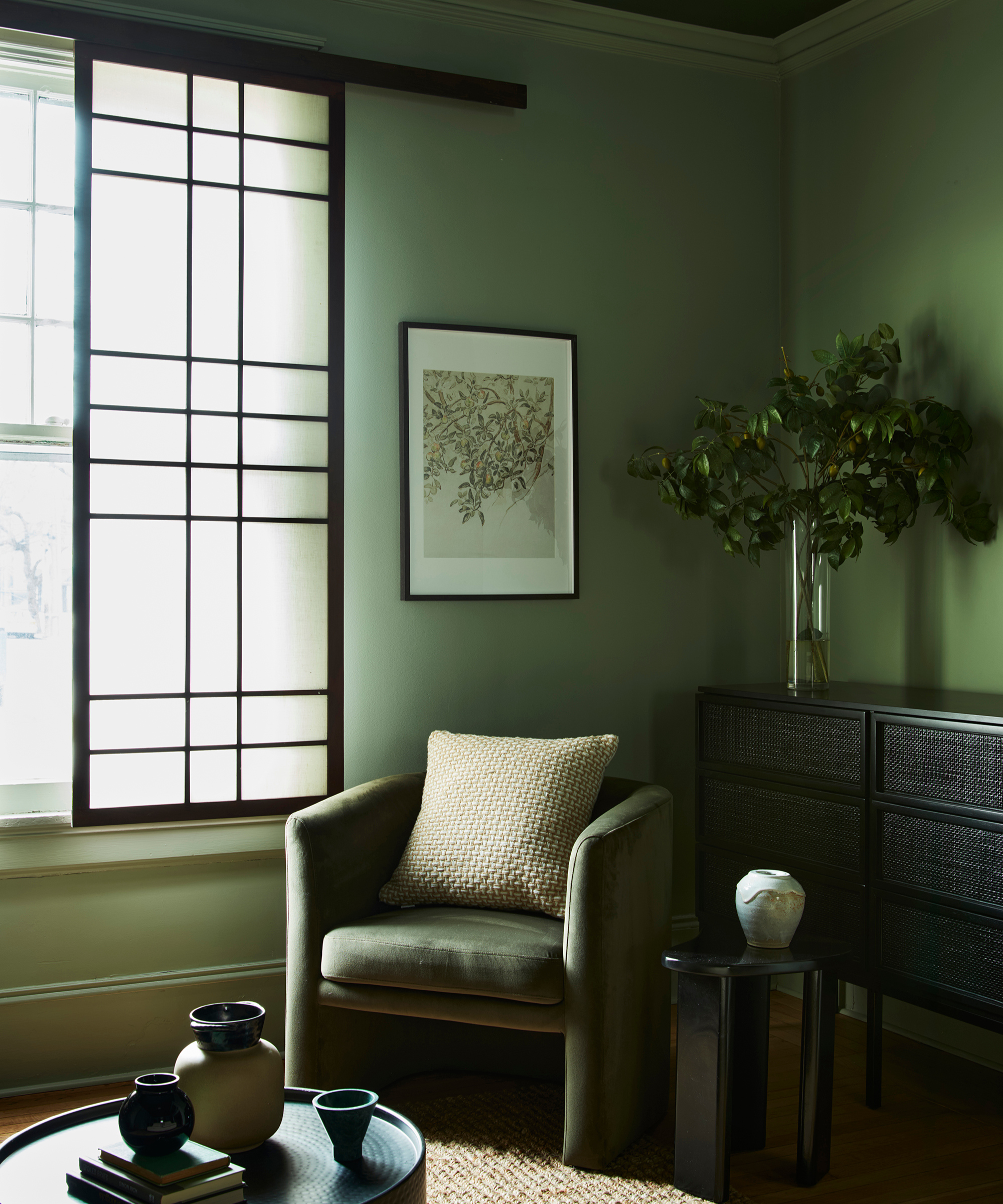 A sage green living room with black panelled windows, a green armchair, and a black console table and coffee table