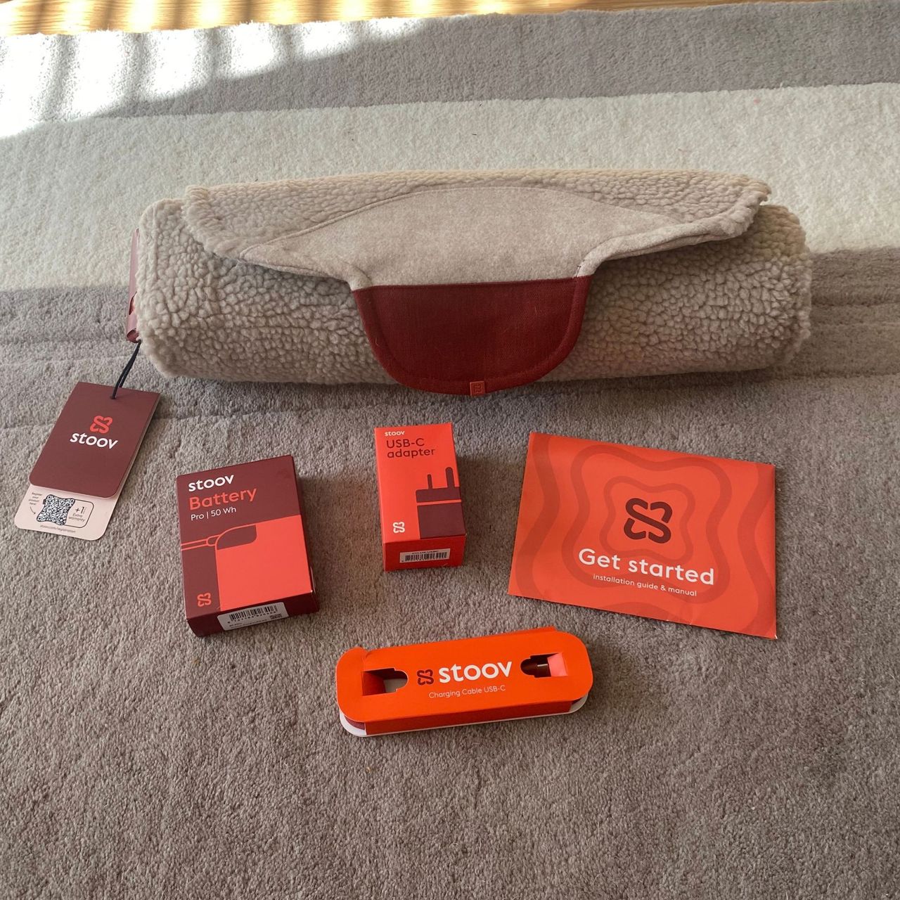 A rolled up Stoov Big Hug3, with the battery, cable and getting started manual on a neutral rug