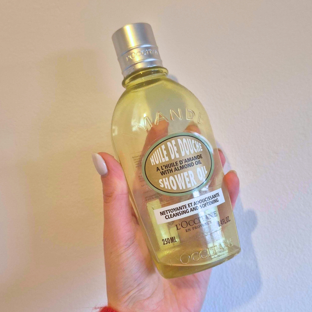 Hand holding empty bottle of L'Occitane Almond Shower Oil against white wall