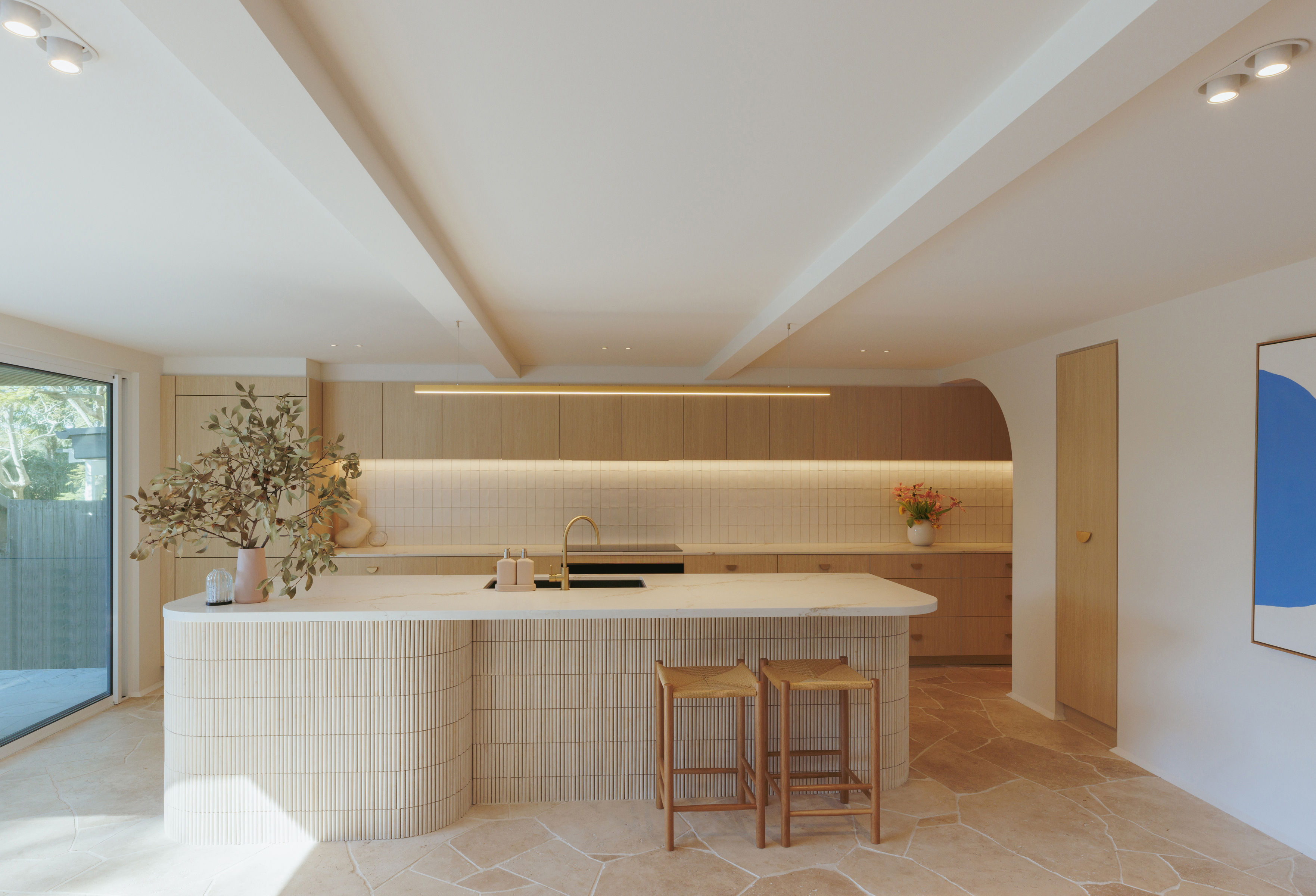 A kitchen in muted tones, with stone flooring and a tiled island in the middle with kitchen stools beside it.