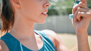 Woman wearing a blue athletic vest is shown looking at her blue asthma inhaler. The background is blurred but it looks like she is standing on a tennis court.