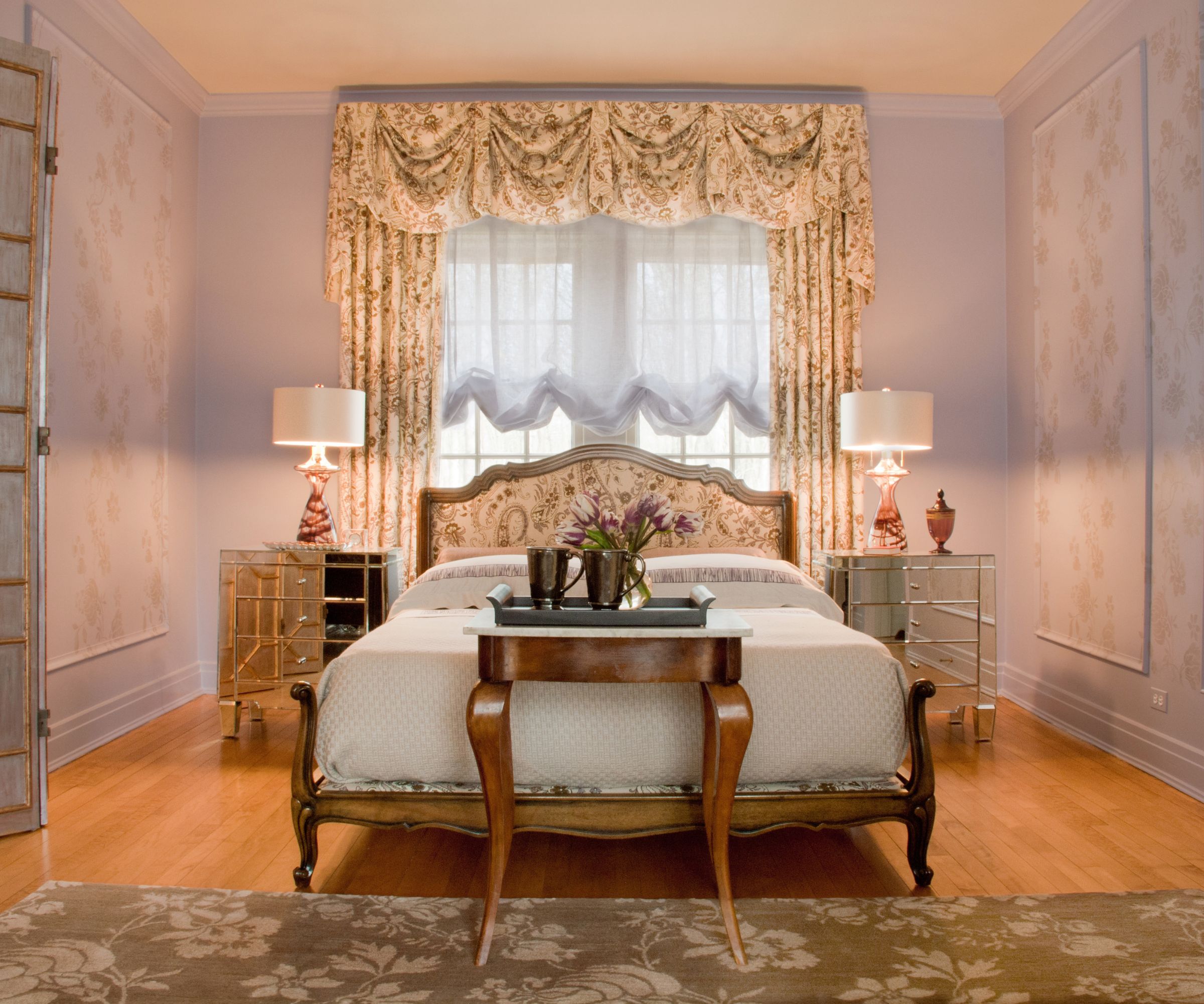 A traditional bedroom with pale lavender walls, wooden flooring, a wooden bedframe with a floral-patterned headboard in front of a window with ruched floral curtains.