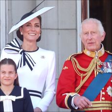 Kate Middleton wears a white dress with navy accents and a matching hat on the Buckingham Palace balcony with King Charles, who wears red military uniform