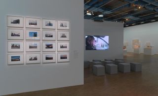 A gallery with a grey wall with many photos on it next to a wall with a screen on it and square grey chairs in front of it.