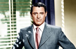 Archie - Cary Grant in a grey suit and red tie stands in front of a window.