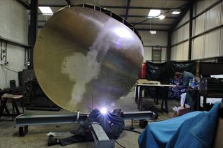 Two men in blue overralls in a work area, working on the installation of a big round metal object