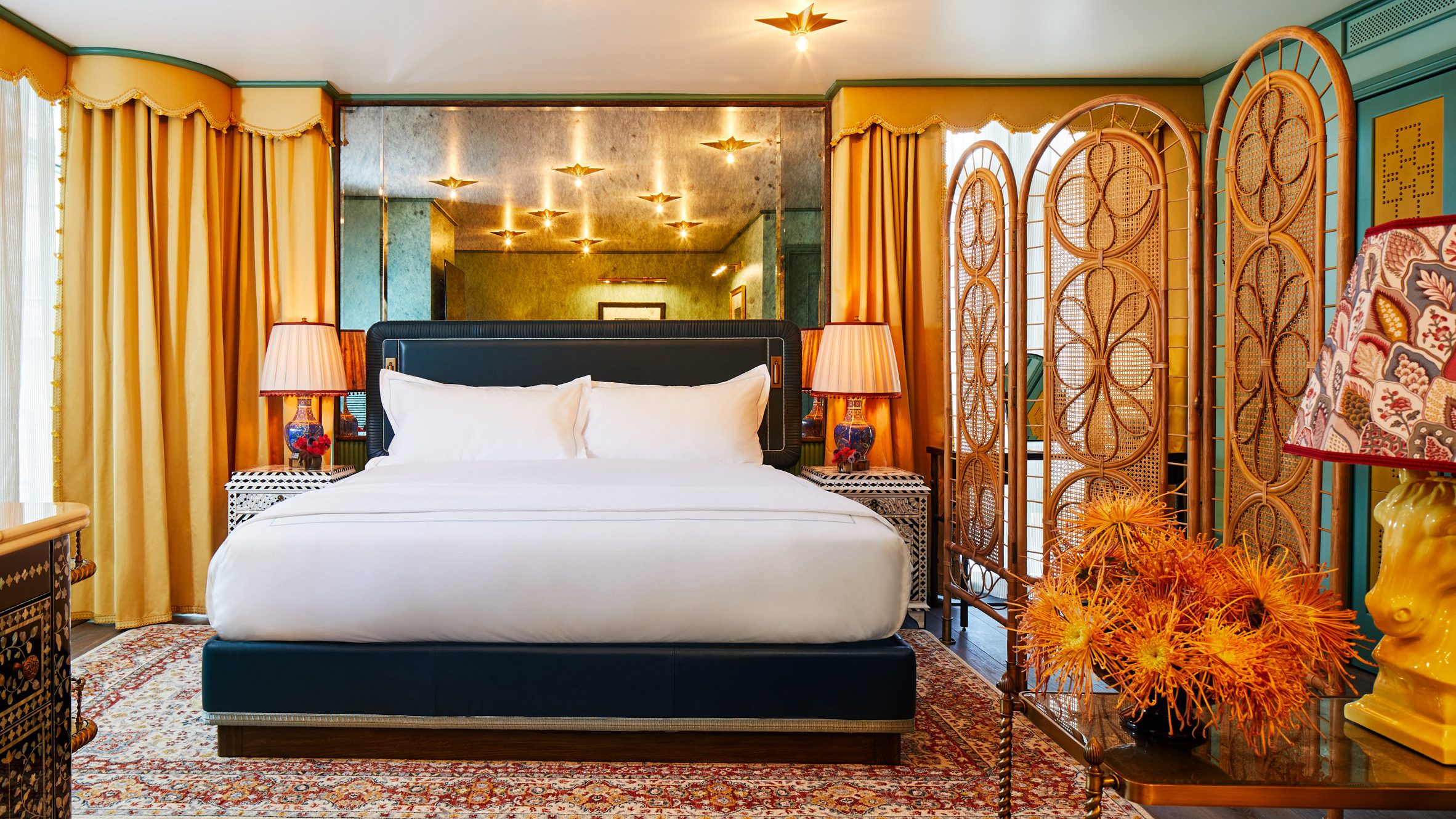 A whimsically decorated, maximalist hotel room is dressed in plush tile, ochre, blue, and terracotta textiles, grand chandeliers and table lamps, and refined upholstery with a nostalgic, retro twist.