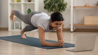 Woman performs plank with leg lift on an exercise mat in her living room, a laptop is open in front of her