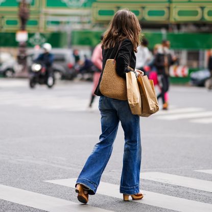 passerby wears a black jacket, a brown straw summer large bag, blue denim flared jeans pants, brown shoes, brown paper shopping bags, on July 03, 2020 in Paris, France. 
