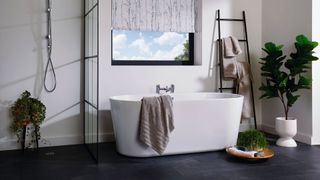 bathroom with black floor and freestanding tub below a window with a towel ladder beside it along with a large houseplant to complete the Japandi bathroom aesthetic