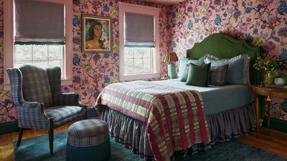 Image of a bedroom with pink chintz wallpaper, and pink trim, and a pink ceiling. There is a purple and blue plaid chair and ottoman, and a bed with a green velvet headboard and blue and pink bedding.