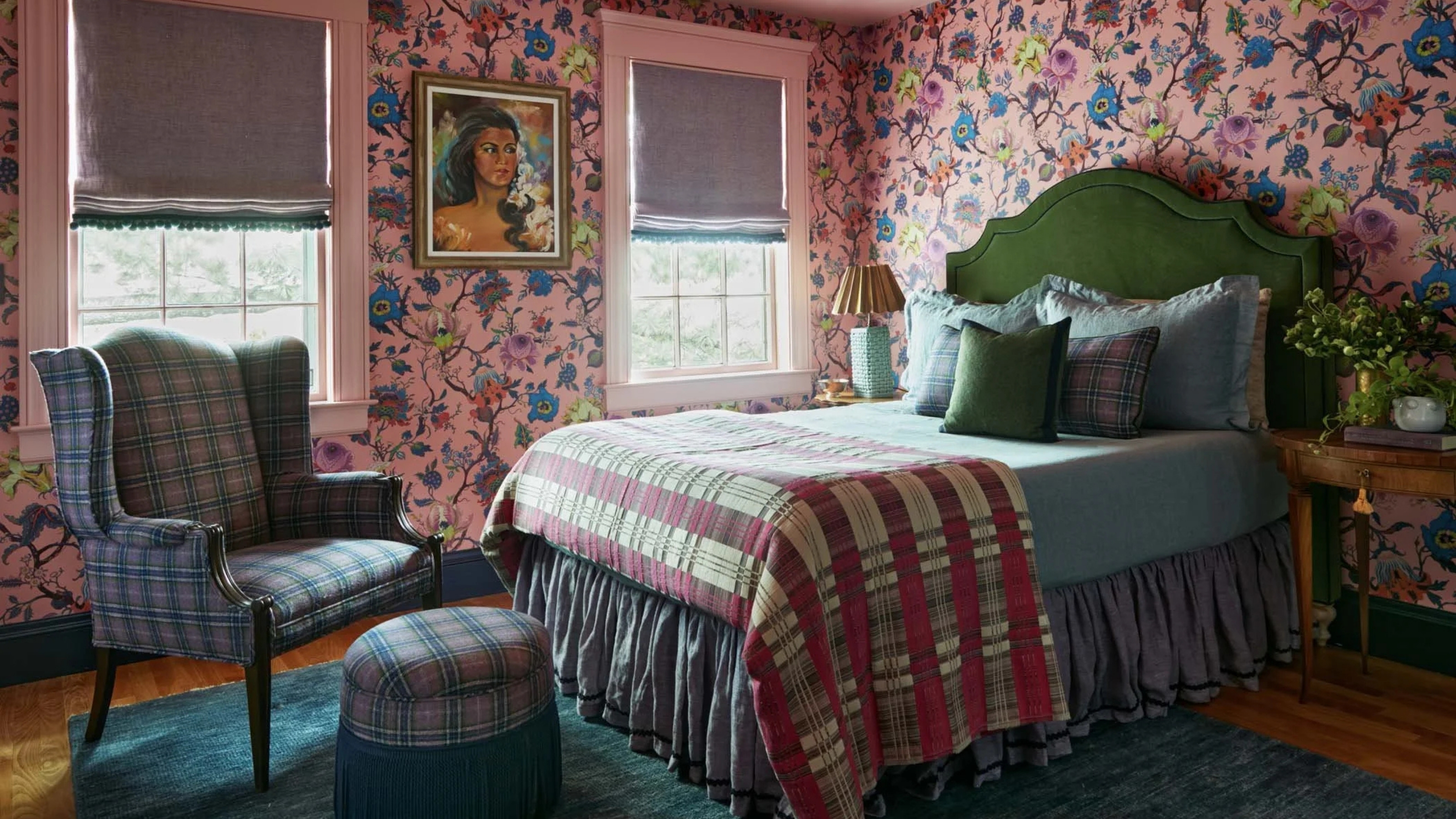 Image of a bedroom with pink chintz wallpaper, and pink trim, and a pink ceiling. There is a purple and blue plaid chair and ottoman, and a bed with a green velvet headboard and blue and pink bedding.