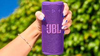 JBL Grip in purple, held in a hand with beautiful nails, in front of a tree 