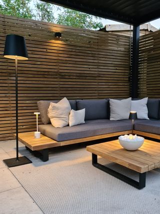 A sheltered backyard area with a grey L sofa and a firepit bowl on a wooden slatted table