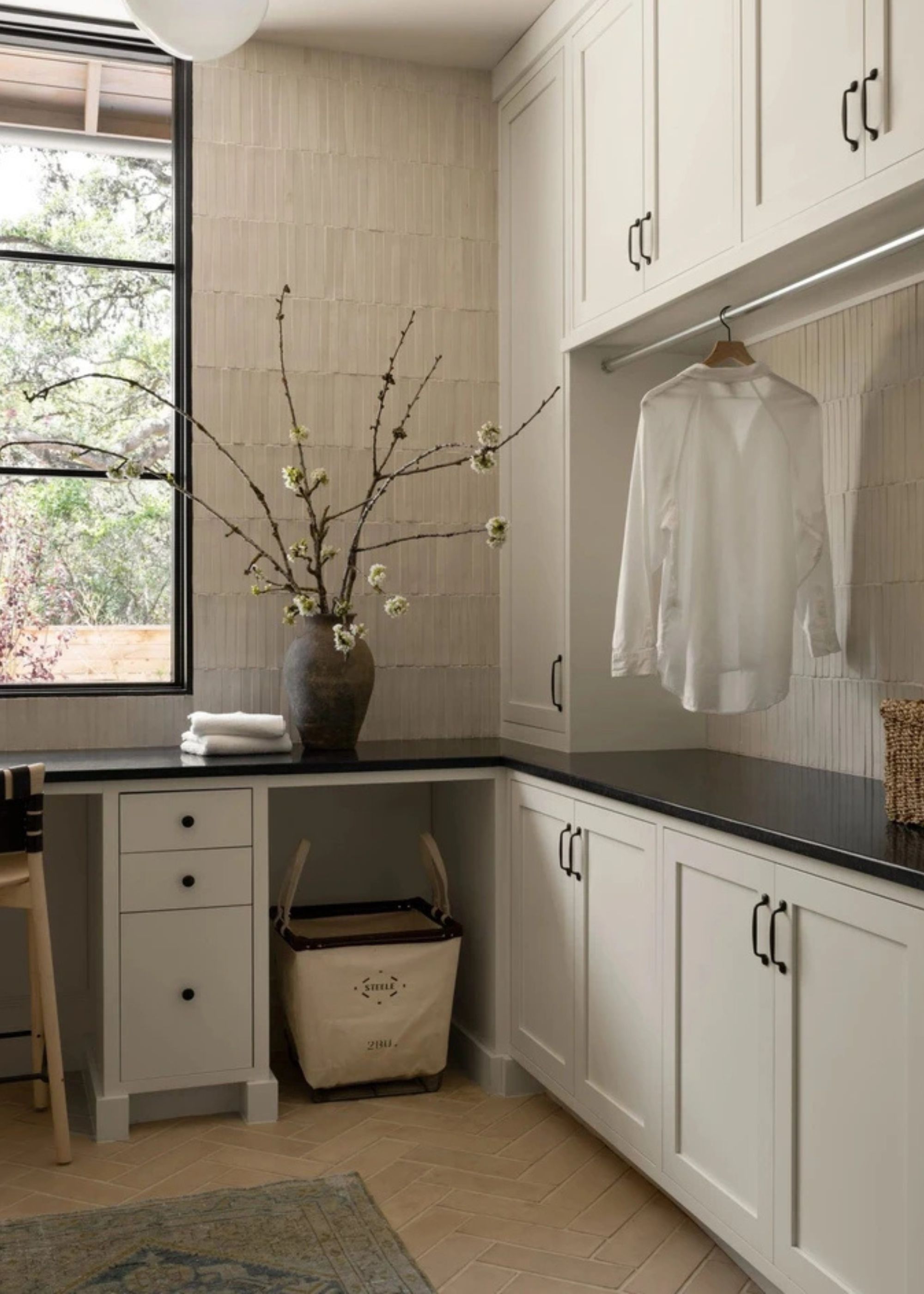 A white utility room with a vase of stems, a laundry basket, a hanging rial, and herringbone floors