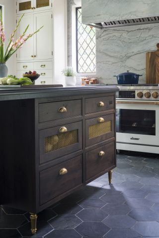 Image of a kitchen that highlights the dark wooden cabinetry with golden hardware on the island. The floors are dark tiles and there is a marble backsplash and hutch.