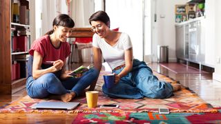 Female couple sat on floor of kitchen on a rug talking with iPad and mugs