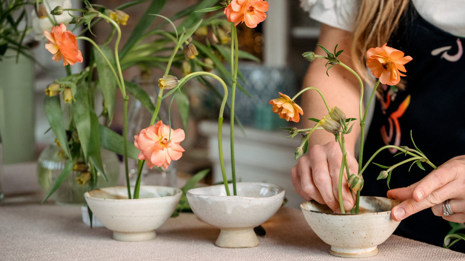 Hands creating a floral design with peach butterfly ranunculus and uva vulpis fritillaries in three ceramic bowls